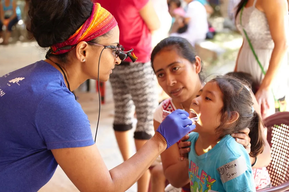 A dental professional wearing a headlamp and gloves examines a young girl's mouth while a woman beside her looks on
